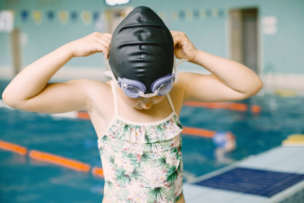 4. Image Alt Text: Young child in swimsuit, goggles, and swim cap standing at poolside ready for their first swimming lesson